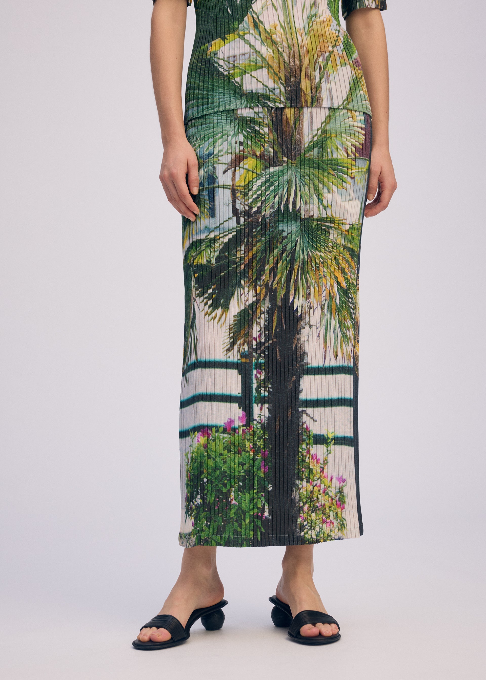 Zoomed view A person wears the ISSEY MIYAKE URBAN JUNGLE PLEATS skirt with a vivid palm and plant print, standing against a plain light background, cropped at the shoulders to show only lower legs and black sandals.