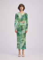 A woman with short curly brown hair wears ISSEY MIYAKE's OBSCURE PLEATS: a green and white ribbed long-sleeve crop top and matching high-waisted, ankle-length moiré-style tie-dye skirt. She stands facing forward in white open-toe heels.