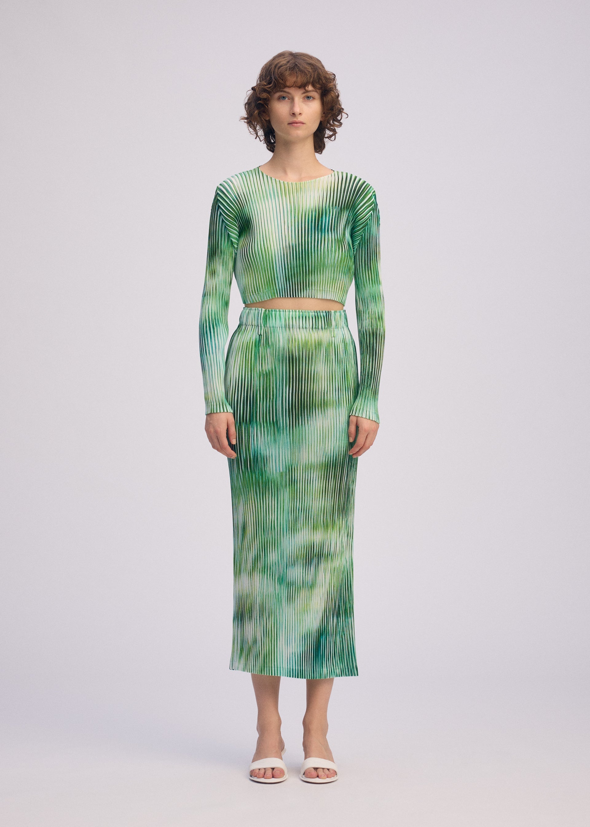 Zoomed view A woman with short curly brown hair wears ISSEY MIYAKE's OBSCURE PLEATS: a green and white ribbed long-sleeve crop top and matching high-waisted, ankle-length moiré-style tie-dye skirt. She stands facing forward in white open-toe heels.