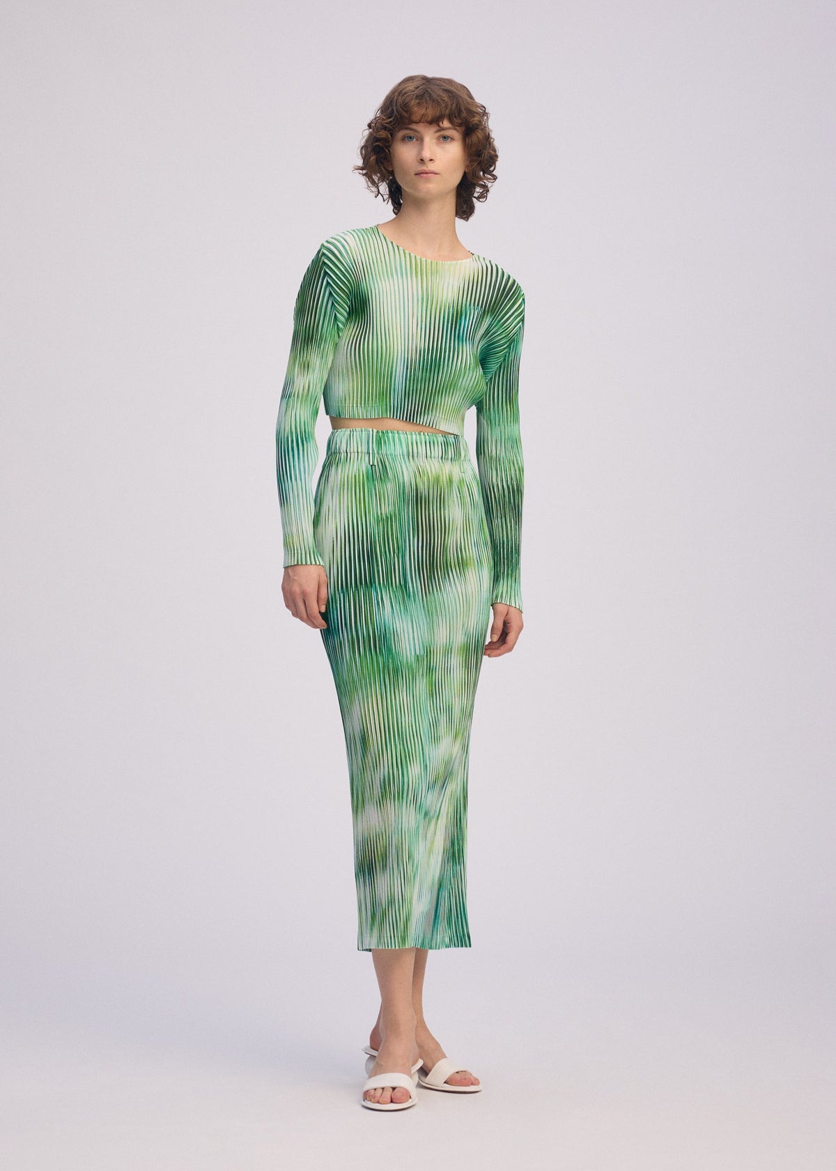 A woman wears the OBSCURE PLEATS dress by ISSEY MIYAKE, featuring green and white pleats. view 7 of 8