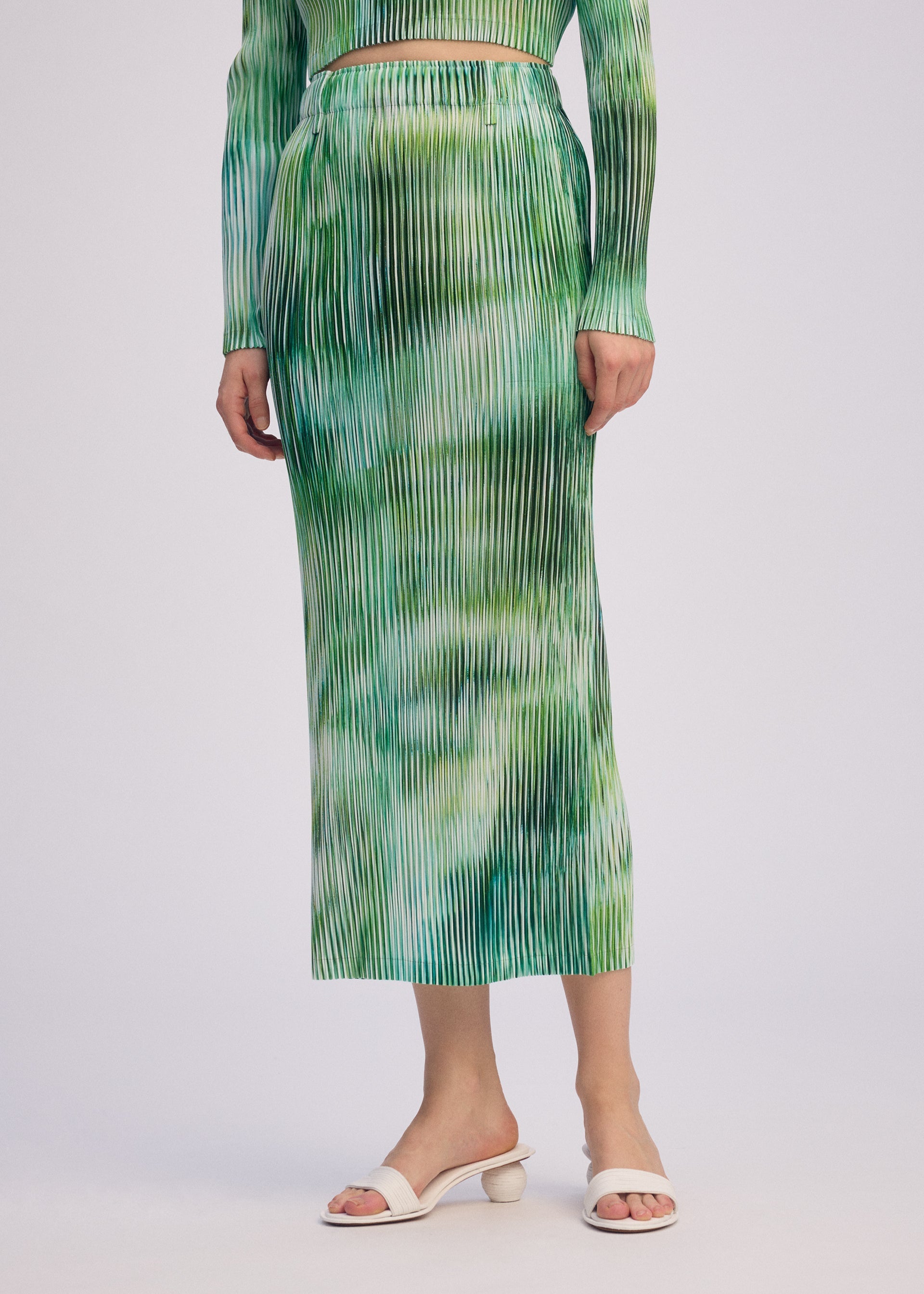 Zoomed view A person models the OBSCURE PLEATS set by ISSEY MIYAKE—green and white striped pleated skirt and matching long-sleeve top—with white open-toe heeled sandals, standing against a plain light background.