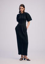 A woman stands against a plain background, wearing the ISSEY MIYAKE LINEN LIKE RIB PLEATS black short-sleeved dress with subtle vertical stripes and a flowing skirt. She has her hands in the dress pockets and wears black sandals.