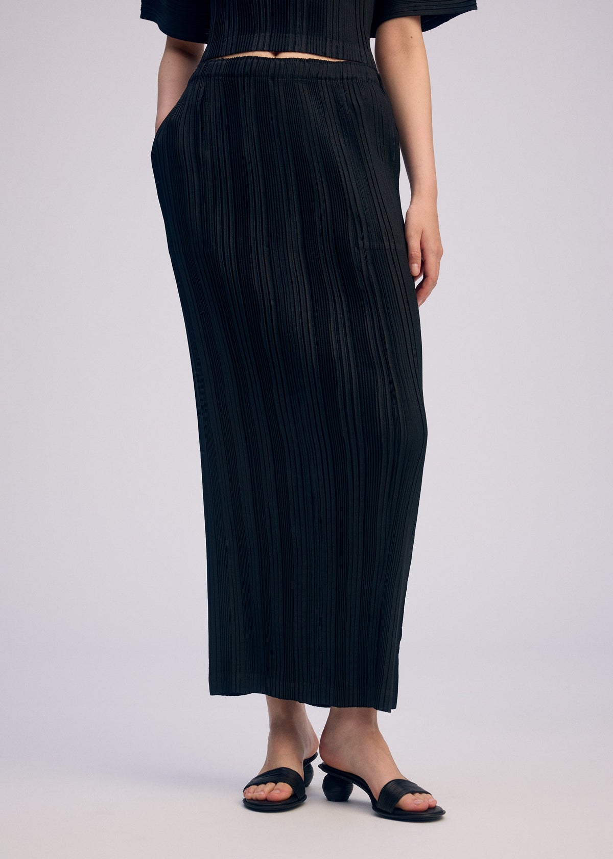 A person wears the ISSEY MIYAKE LINEN LIKE RIB PLEATS set—long pleated skirt with matching top—and black heeled sandals, posing with one hand in a pocket against a plain light background. view 8 of 9