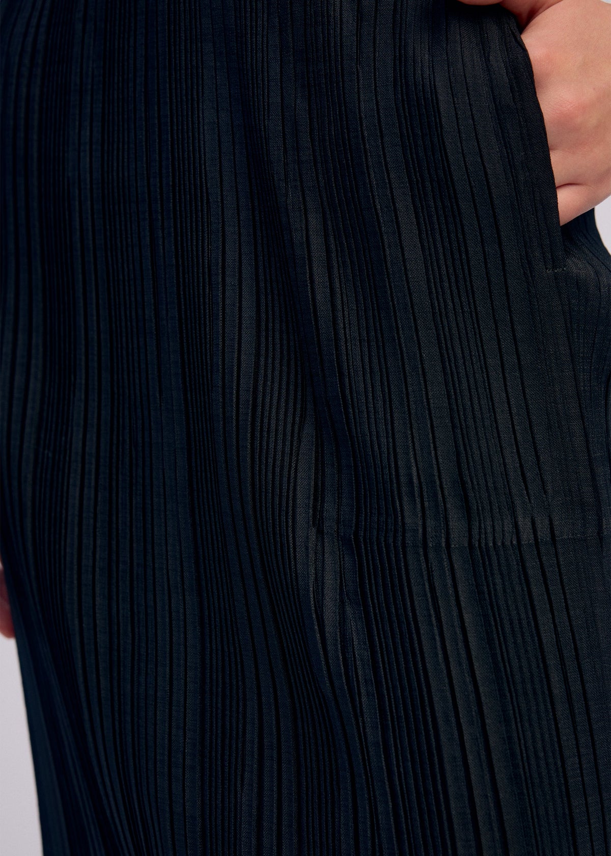 Close-up of a person wearing ISSEY MIYAKE’s LINEN LIKE RIB PLEATS skirt, showing their hand in a side pocket and highlighting the textured vertical lines of the dark, pleated fabric. view 9 of 9