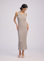 A woman stands against a plain background wearing the ISSEY MIYAKE WAVELET ENSEMBLE sleeveless jacquard stretch maxi dress in light gray, paired with white open-toe sandals. She looks down, gently touching the sides of her dress.