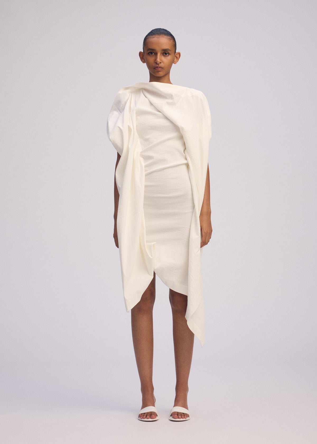 A person stands facing forward in ISSEY MIYAKE's CONCEALED dress—an off-white, textured cotton piece with gathered fabric and an uneven hem—paired with white sandals against a plain light gray background. view 4 of 5