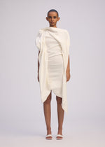 A person stands facing forward in ISSEY MIYAKE's CONCEALED dress—an off-white, textured cotton piece with gathered fabric and an uneven hem—paired with white sandals against a plain light gray background.