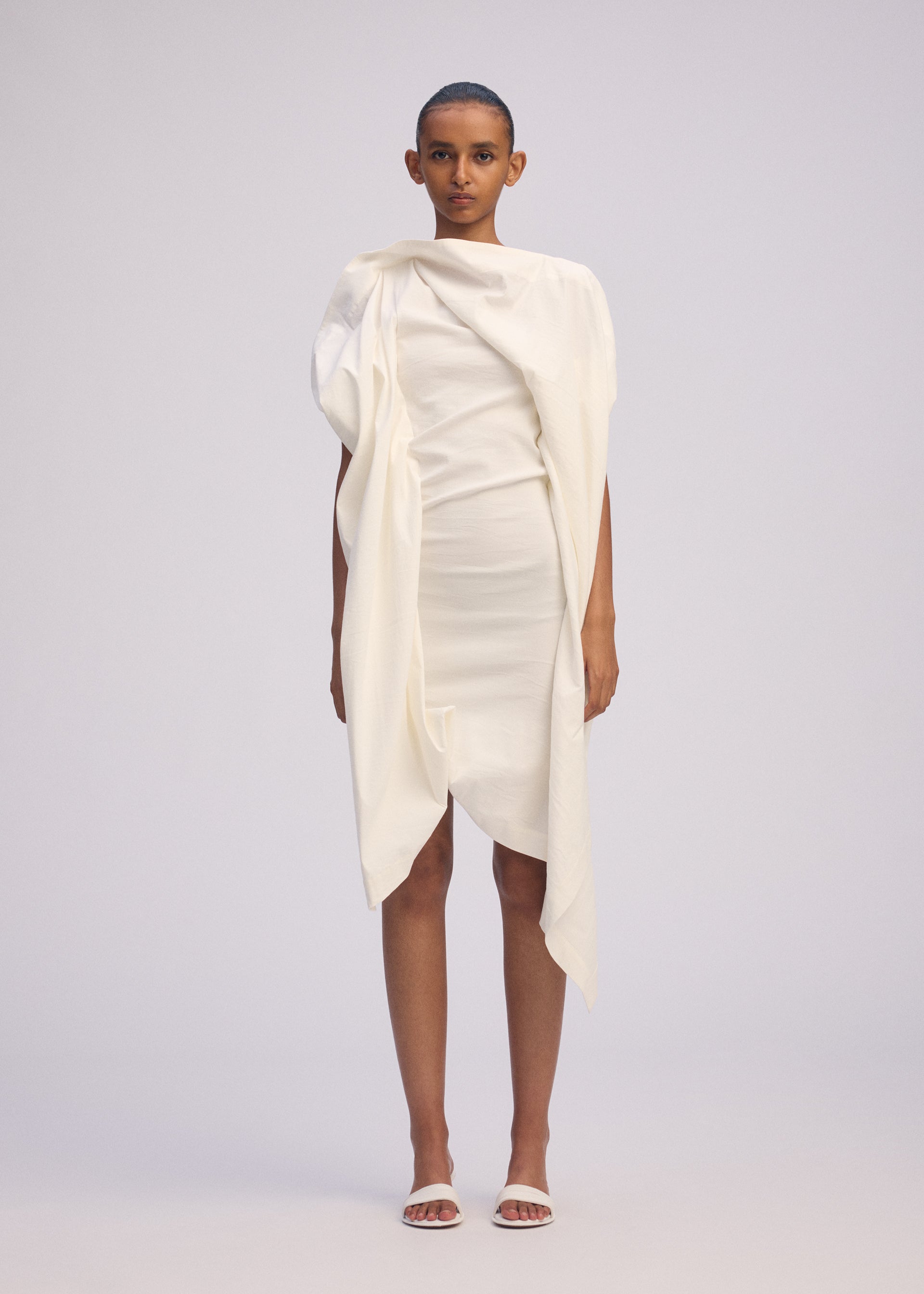 Zoomed view A person stands facing forward in ISSEY MIYAKE's CONCEALED dress—an off-white, textured cotton piece with gathered fabric and an uneven hem—paired with white sandals against a plain light gray background.