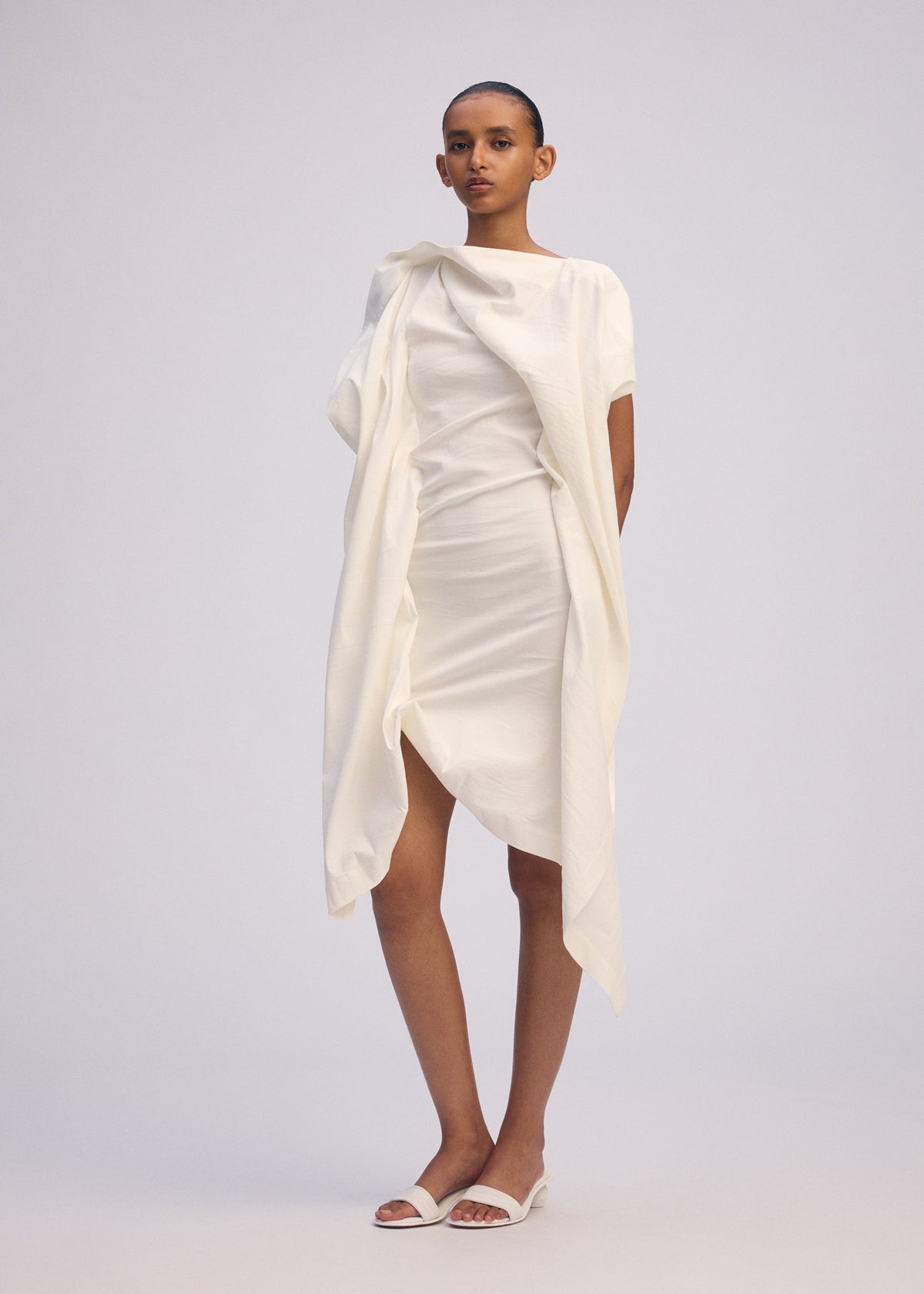 A person with short hair and a neutral expression stands against a plain background wearing ISSEY MIYAKE's CONCEALED dress—an asymmetrical, draped design in Japanese Washi with short sleeves—paired with white open-toe sandals. view 5 of 5
