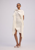 A person with short hair and a neutral expression stands against a plain background wearing ISSEY MIYAKE's CONCEALED dress—an asymmetrical, draped design in Japanese Washi with short sleeves—paired with white open-toe sandals.