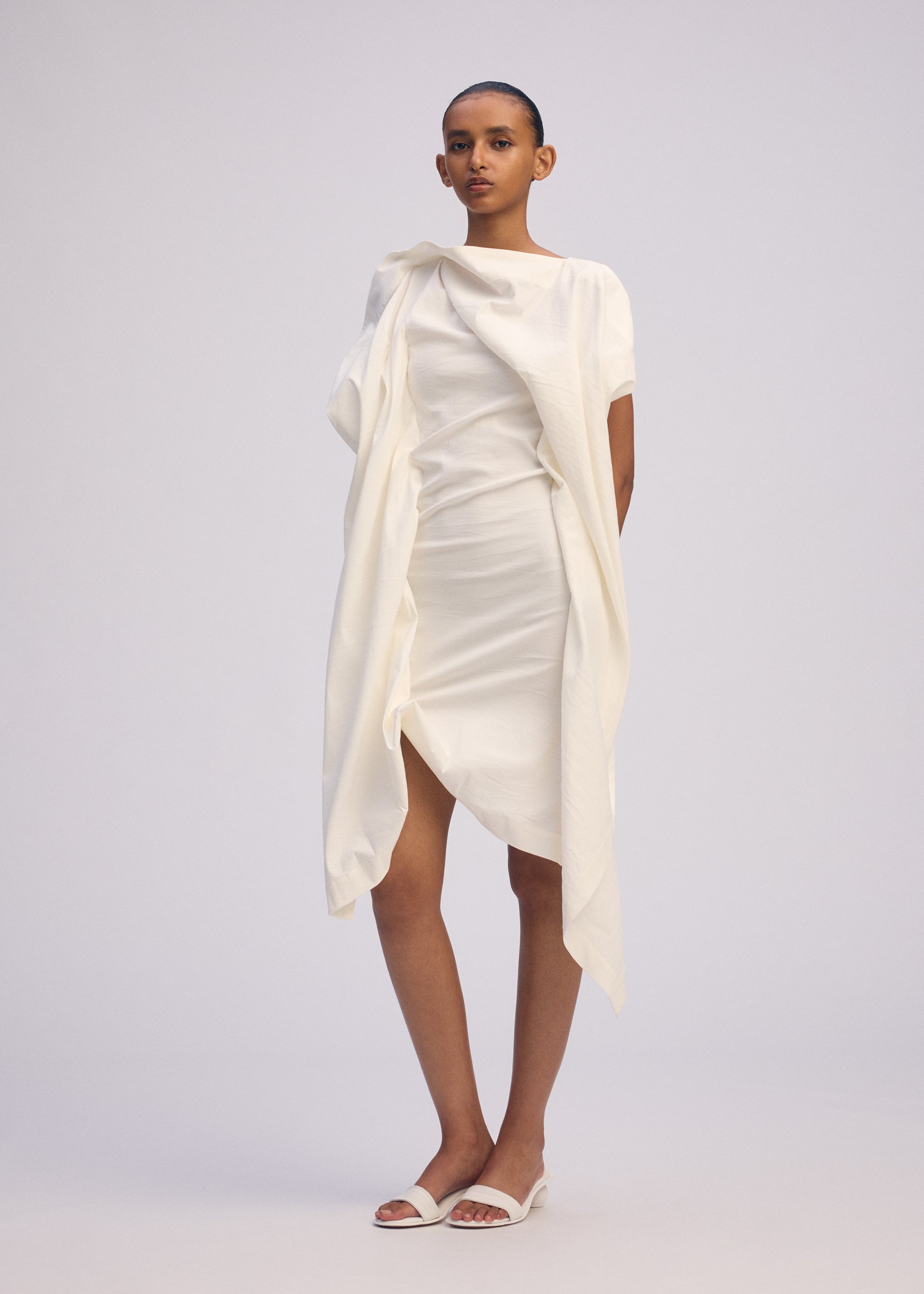 Zoomed view A person with short hair and a neutral expression stands against a plain background wearing ISSEY MIYAKE's CONCEALED dress—an asymmetrical, draped design in Japanese Washi with short sleeves—paired with white open-toe sandals.