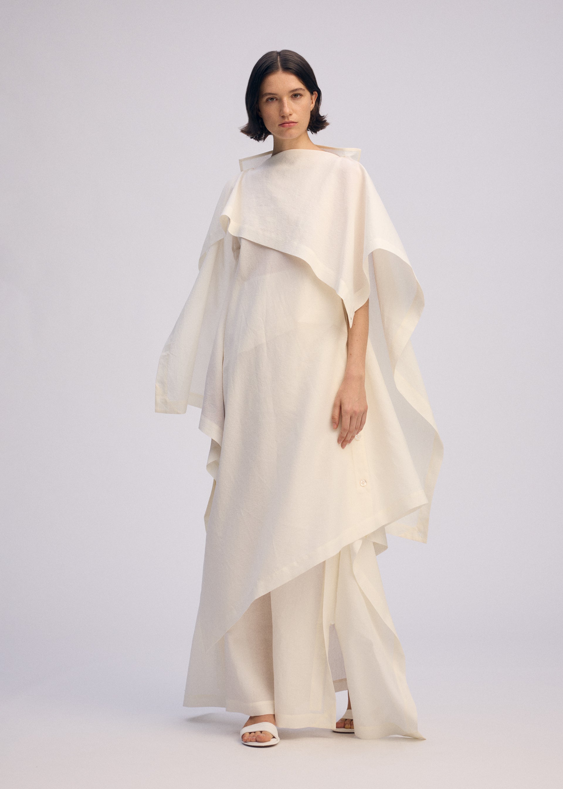 Zoomed view A woman with short dark hair wears the ALL SQUARES ensemble by ISSEY MIYAKE, crafted from ultra-fine polyester with wide, flowing sleeves and pants. She poses in white sandals against a plain, light background.