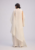 A person with short dark hair faces away, wearing ISSEY MIYAKE’s ALL SQUARES—an elegant, loose-fitting white outfit with asymmetrical draping and wide pants inspired by Japanese Washi paper, set against a plain light background.