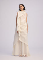 A woman with short dark hair and a neutral expression wears the ISSEY MIYAKE ALL SQUARES sleeveless ivory dress, featuring layered draped fabric inspired by Japanese Washi paper. She stands against a plain background in white sandals.