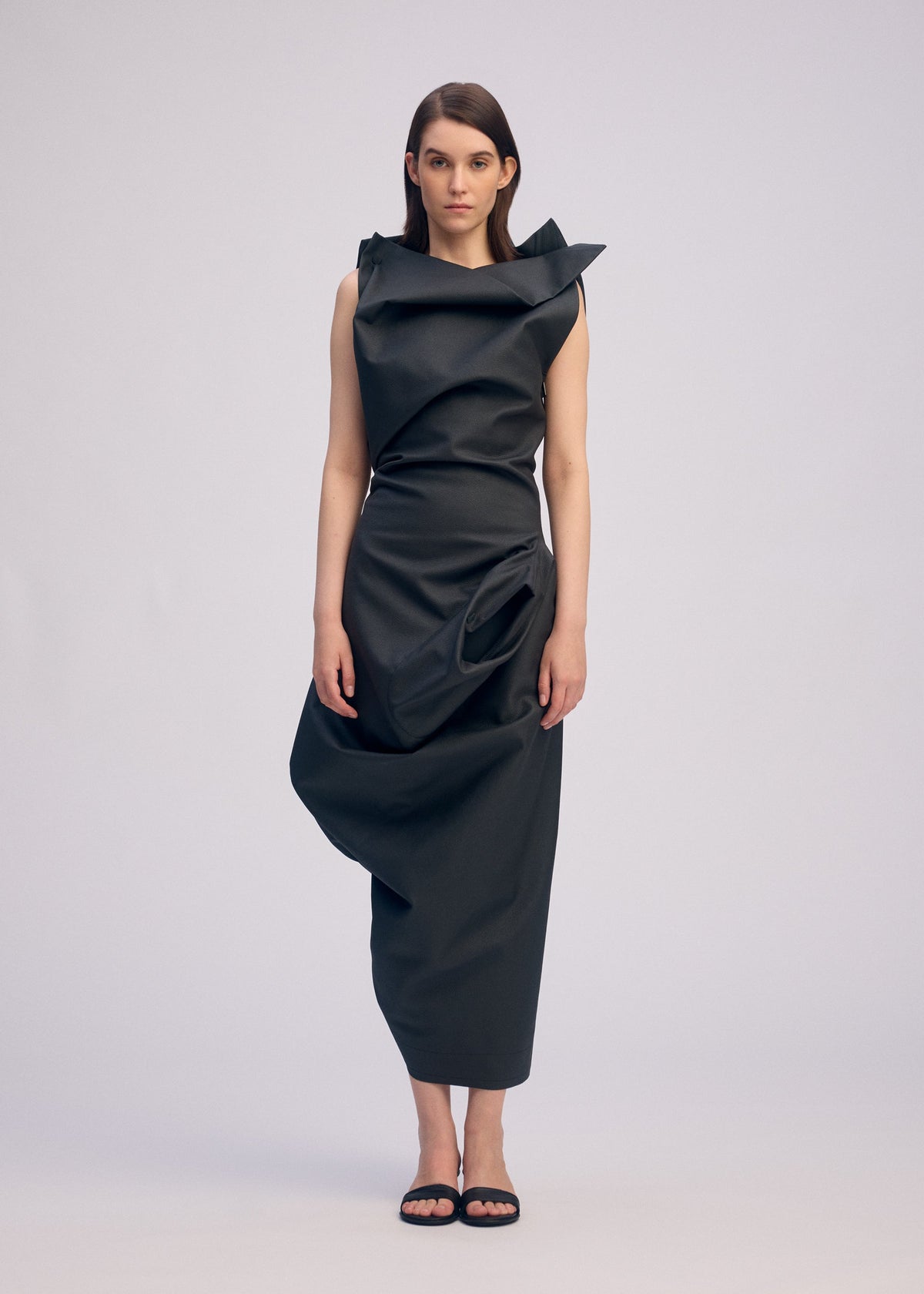 A woman faces forward in the ISSEY MIYAKE PEU FORM³ black artificial leather dress with asymmetrical folds from the Autumn Winter 2025/26 Collection. She has straight brown hair, wears black sandals, and stands against a plain light background. view 5 of 6