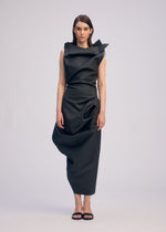 A woman faces forward in the ISSEY MIYAKE PEU FORM³ black artificial leather dress with asymmetrical folds from the Autumn Winter 2025/26 Collection. She has straight brown hair, wears black sandals, and stands against a plain light background.