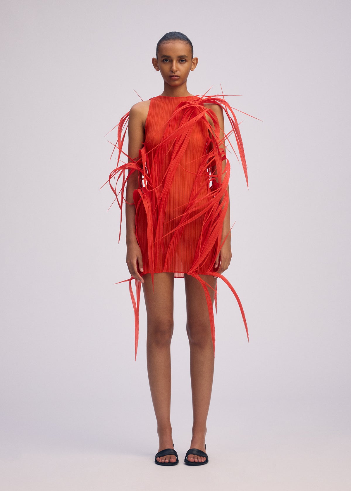A person poses against a plain background wearing ISSEY MIYAKE's URBAN JUNGLE: a striking red mini dress with dramatic, spiky 3D accents and bold textured details extending outward, paired with black sandals. view 5 of 7