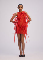 A model in ISSEY MIYAKE’s URBAN JUNGLE mini dress—vivid orange with sculptural, ribbon-like extensions and dynamic flow—stands against a plain light background, sporting short hair and black sandals.