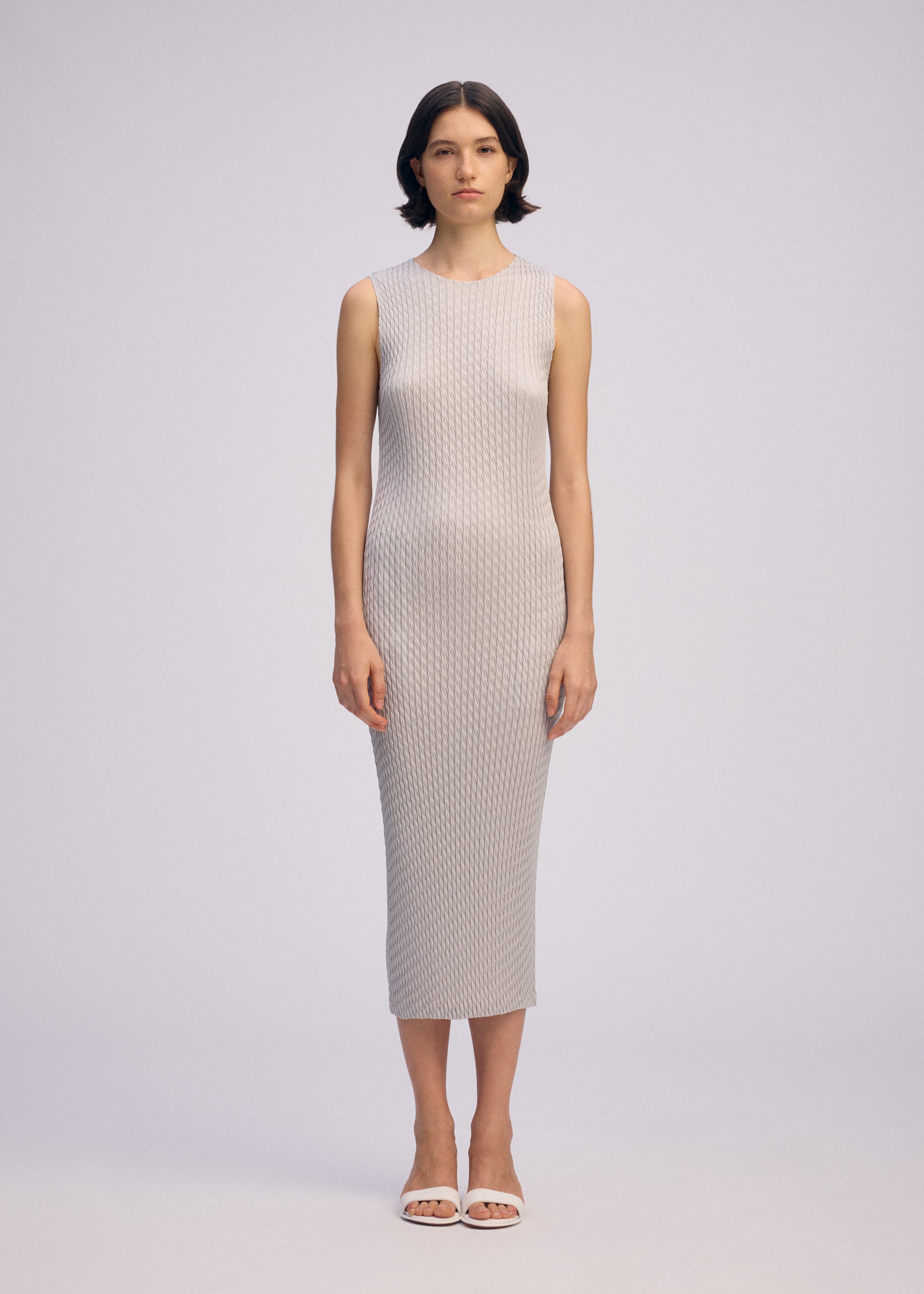 Zoomed view A woman stands facing forward, wearing ISSEY MIYAKE's SLEEK PLEATS round-neck sleeveless light gray ankle-length polyester dress with a subtle textured pattern and white open-toe sandals, against a plain light background.