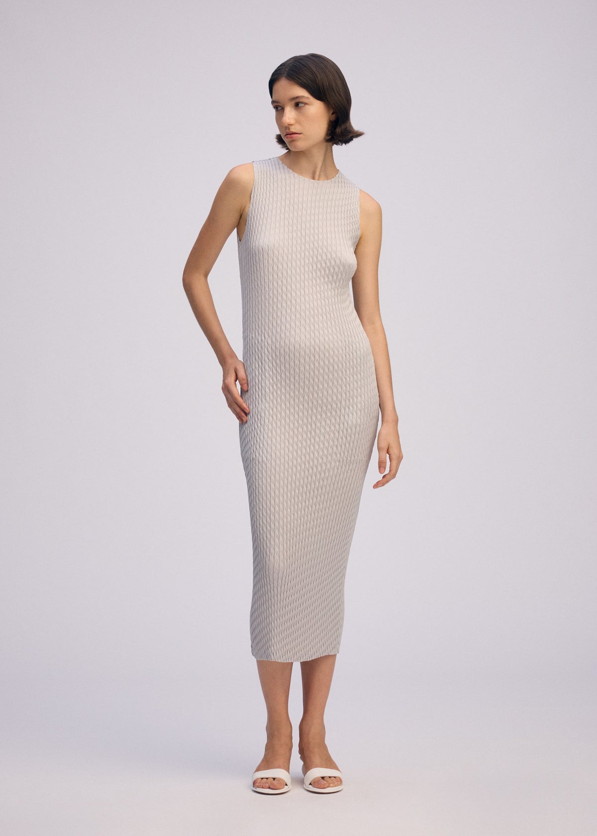 A woman poses against a light background wearing the ISSEY MIYAKE SLEEK PLEATS round-neck sleeveless, form-fitting textured light gray midi dress and white open-toe sandals, with one hand by her side and the other touching her hip. view 6 of 6