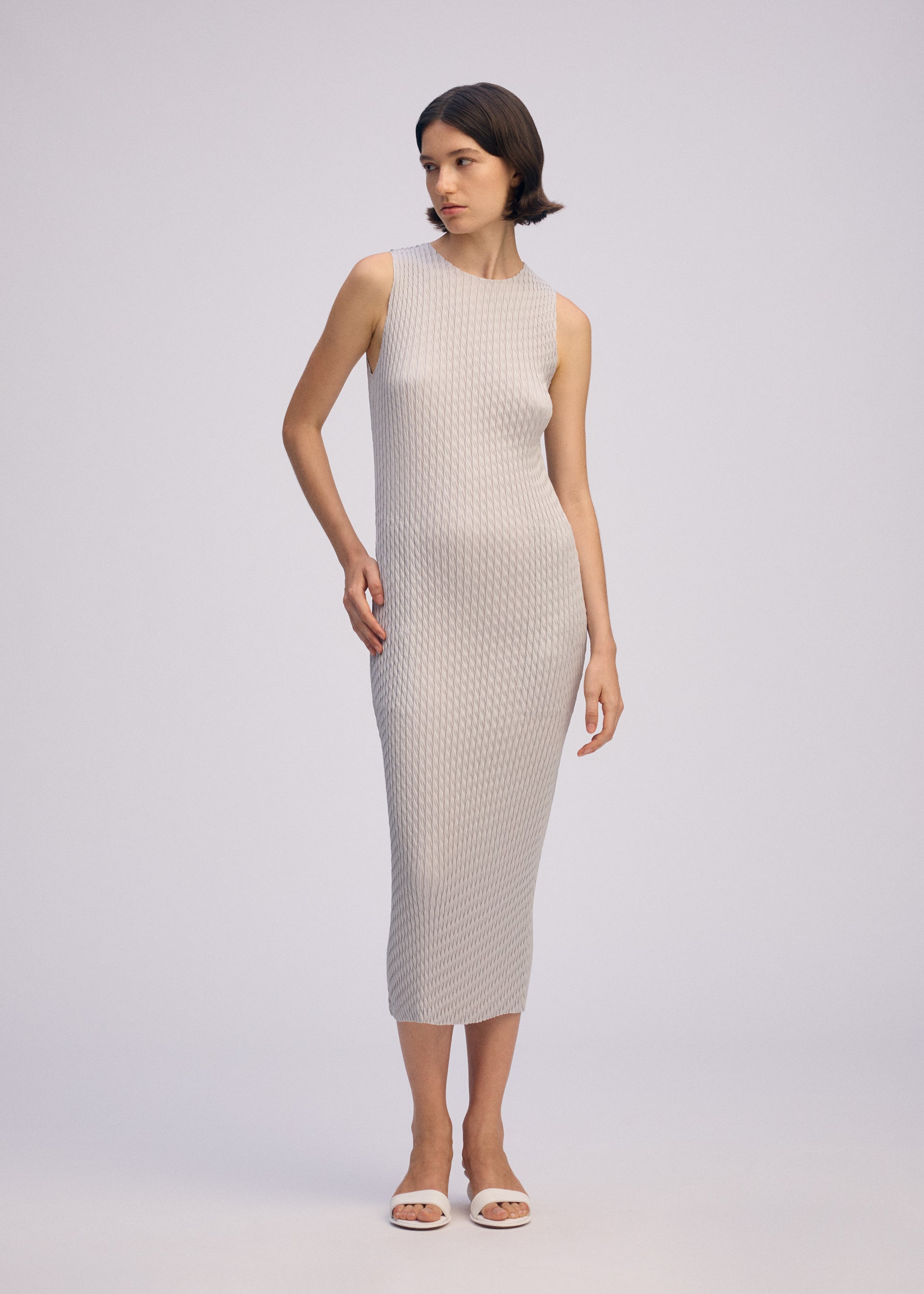 Zoomed view A woman poses against a light background wearing the ISSEY MIYAKE SLEEK PLEATS round-neck sleeveless, form-fitting textured light gray midi dress and white open-toe sandals, with one hand by her side and the other touching her hip.