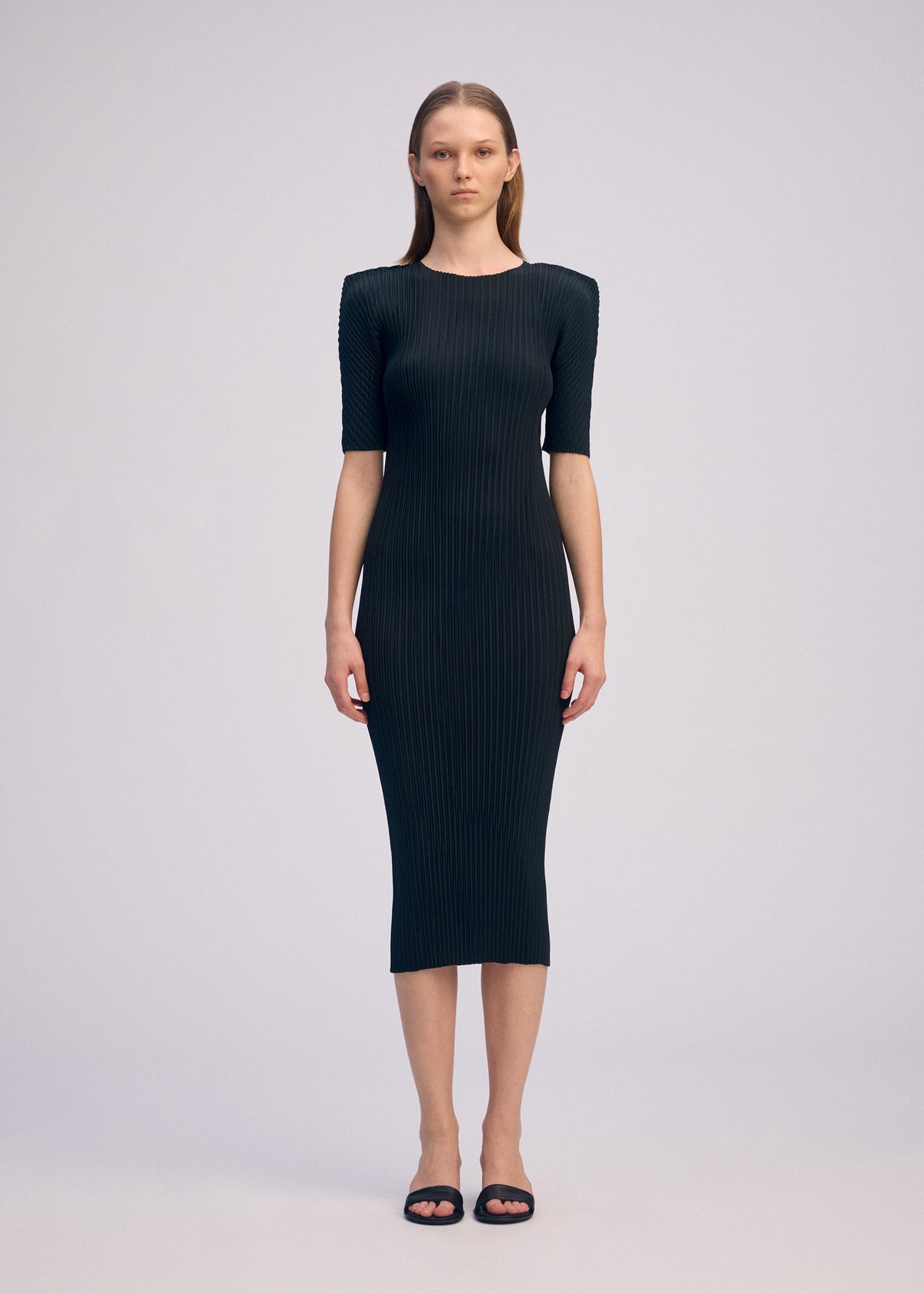 A woman faces forward in a fitted, ribbed black BOX SHOULDER PLEATS dress by ISSEY MIYAKE with short sleeves and a round neck, paired with black sandals. The background is plain and light-colored. view 5 of 6