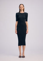 A woman faces forward in a fitted, ribbed black BOX SHOULDER PLEATS dress by ISSEY MIYAKE with short sleeves and a round neck, paired with black sandals. The background is plain and light-colored.