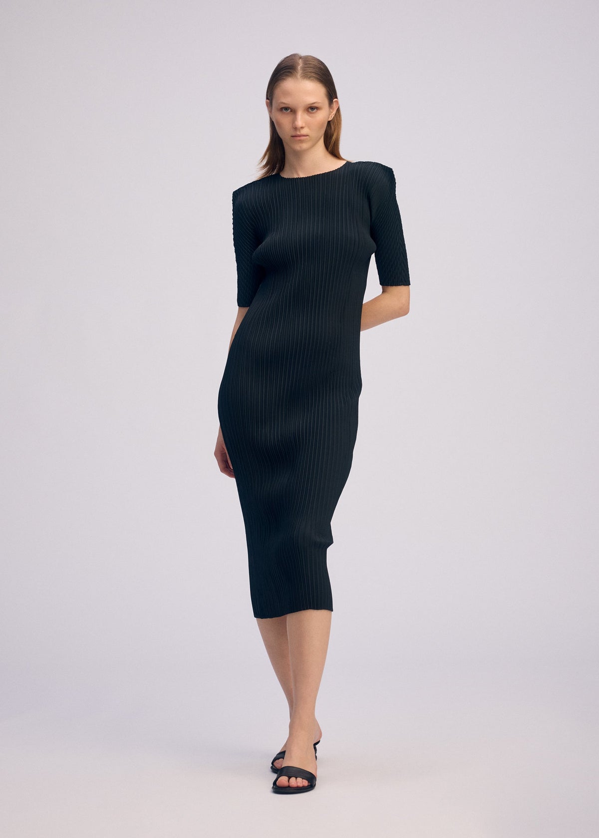 A woman with straight, shoulder-length hair wears ISSEY MIYAKE's BOX SHOULDER PLEATS: a fitted, ribbed black midi dress with elbow-length sleeves and a box shoulder silhouette, paired with black heeled sandals against a plain background. view 6 of 6