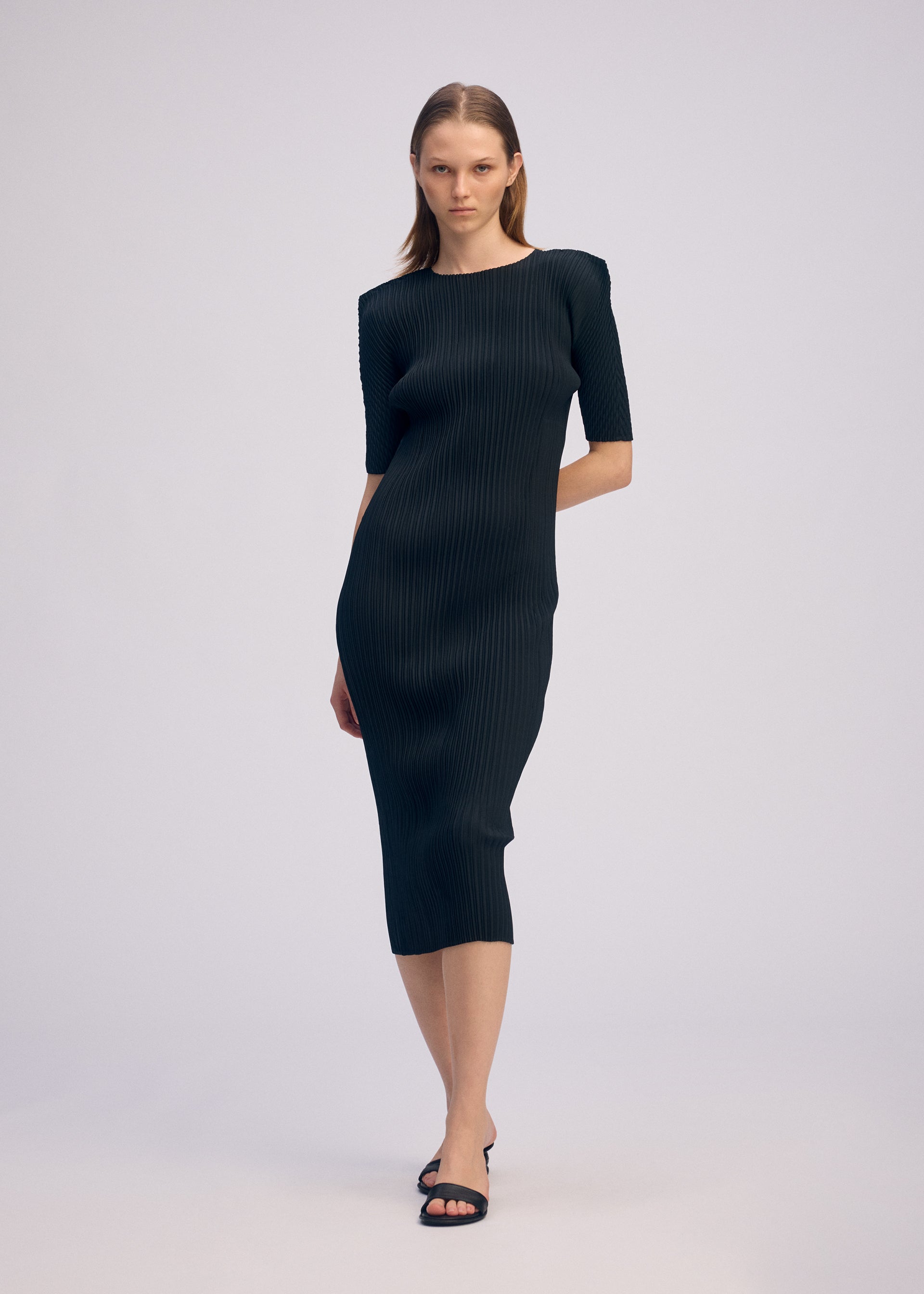 Zoomed view A woman with straight, shoulder-length hair wears ISSEY MIYAKE's BOX SHOULDER PLEATS: a fitted, ribbed black midi dress with elbow-length sleeves and a box shoulder silhouette, paired with black heeled sandals against a plain background.