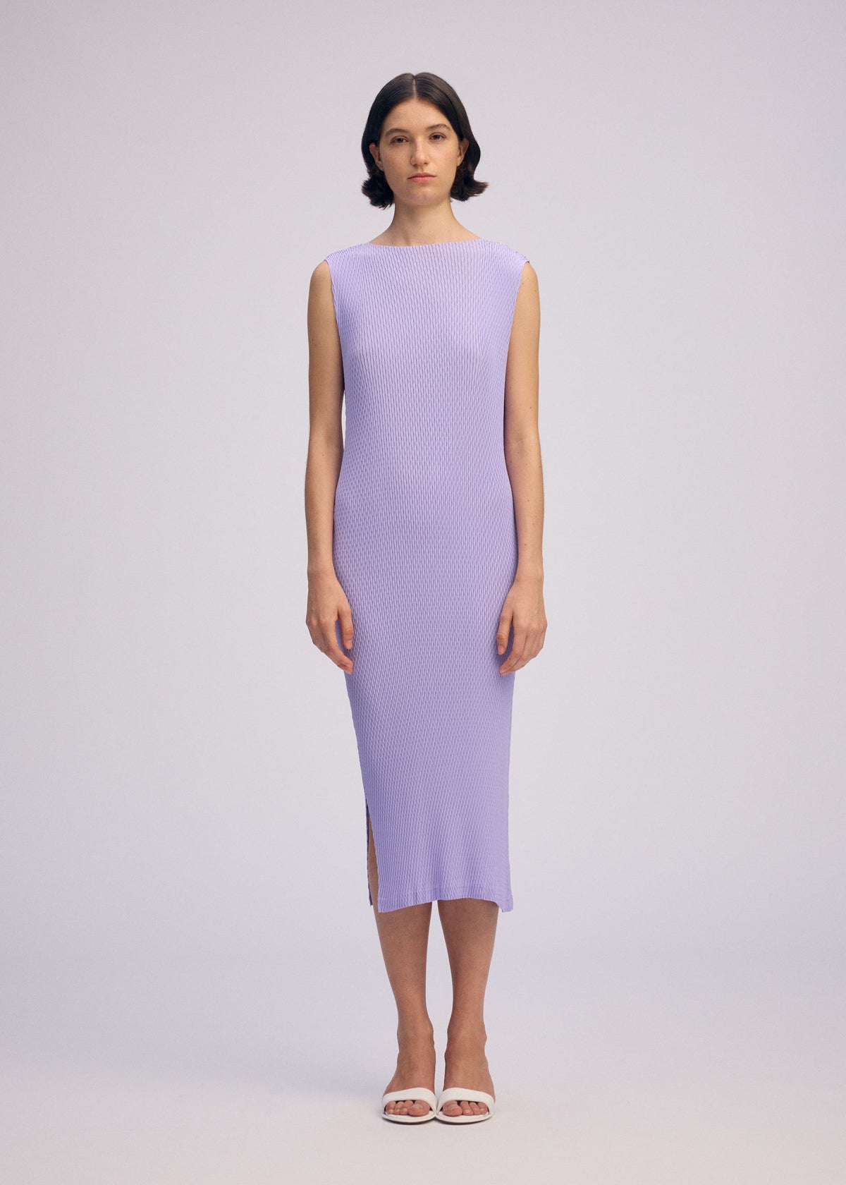 A woman with short dark hair faces forward in the ISSEY MIYAKE RHOMBUS PLEATS dress, a light purple, ankle-length, ribbed sleeveless style with a round neck and side slit. She pairs it with white sandals against a plain light background. view 4 of 6