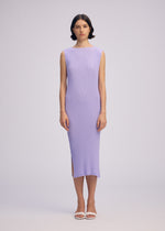 A woman with short dark hair faces forward in the ISSEY MIYAKE RHOMBUS PLEATS dress, a light purple, ankle-length, ribbed sleeveless style with a round neck and side slit. She pairs it with white sandals against a plain light background.