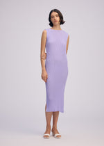 A woman stands against a plain background wearing the ISSEY MIYAKE RHOMBUS PLEATS, a light purple, ankle-length, round-neck sleeveless dress with a sleek silhouette. She pairs it with white sandals and has short dark hair in loose waves.