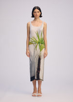 A woman with short dark hair and white sandals stands against a plain light background, wearing ISSEY MIYAKE's URBAN JUNGLE PLEATS: a sleeveless, ankle-length ribbed dress featuring a bold green palm tree print on the front.