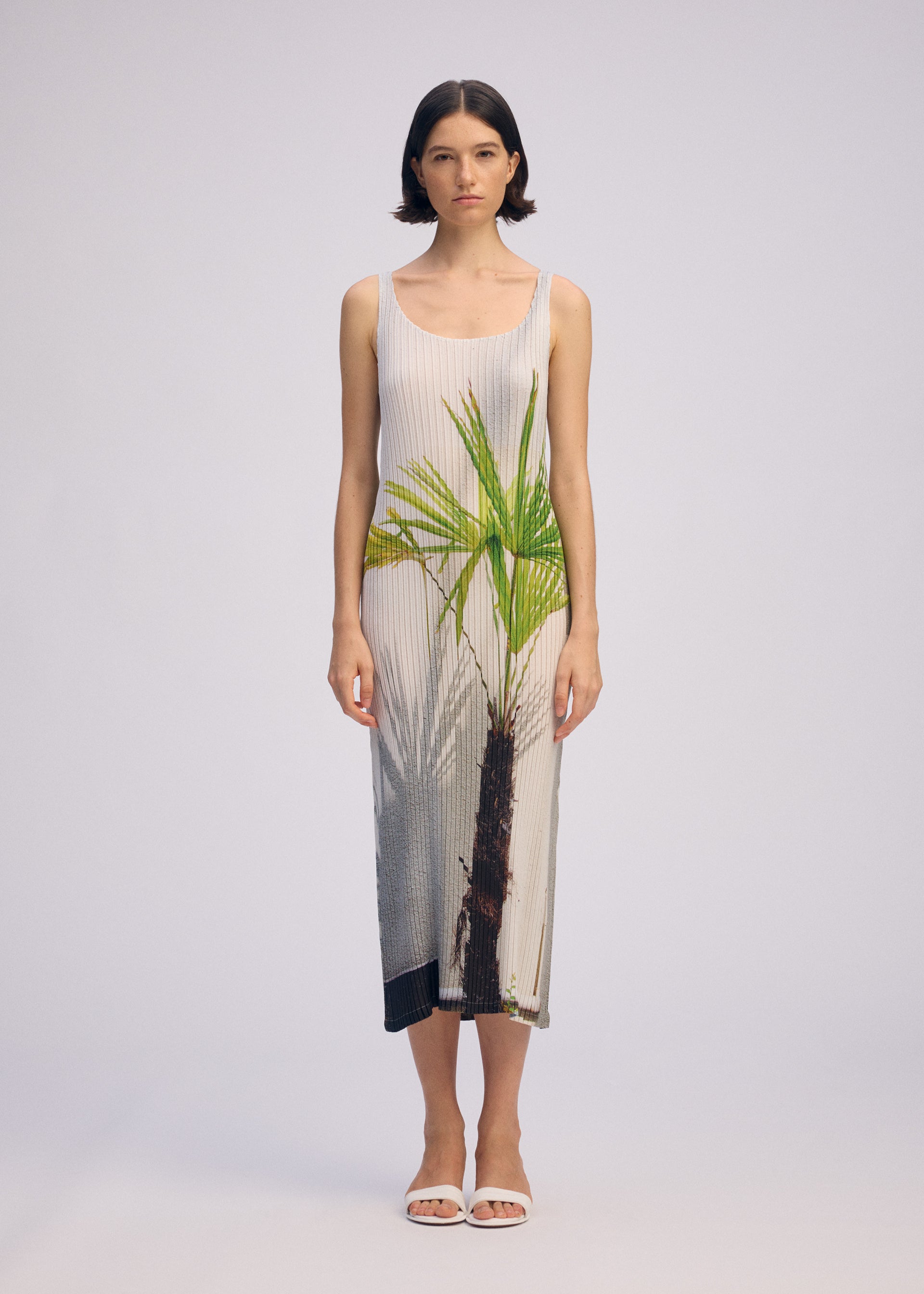 Zoomed view A woman with short dark hair and white sandals stands against a plain light background, wearing ISSEY MIYAKE's URBAN JUNGLE PLEATS: a sleeveless, ankle-length ribbed dress featuring a bold green palm tree print on the front.
