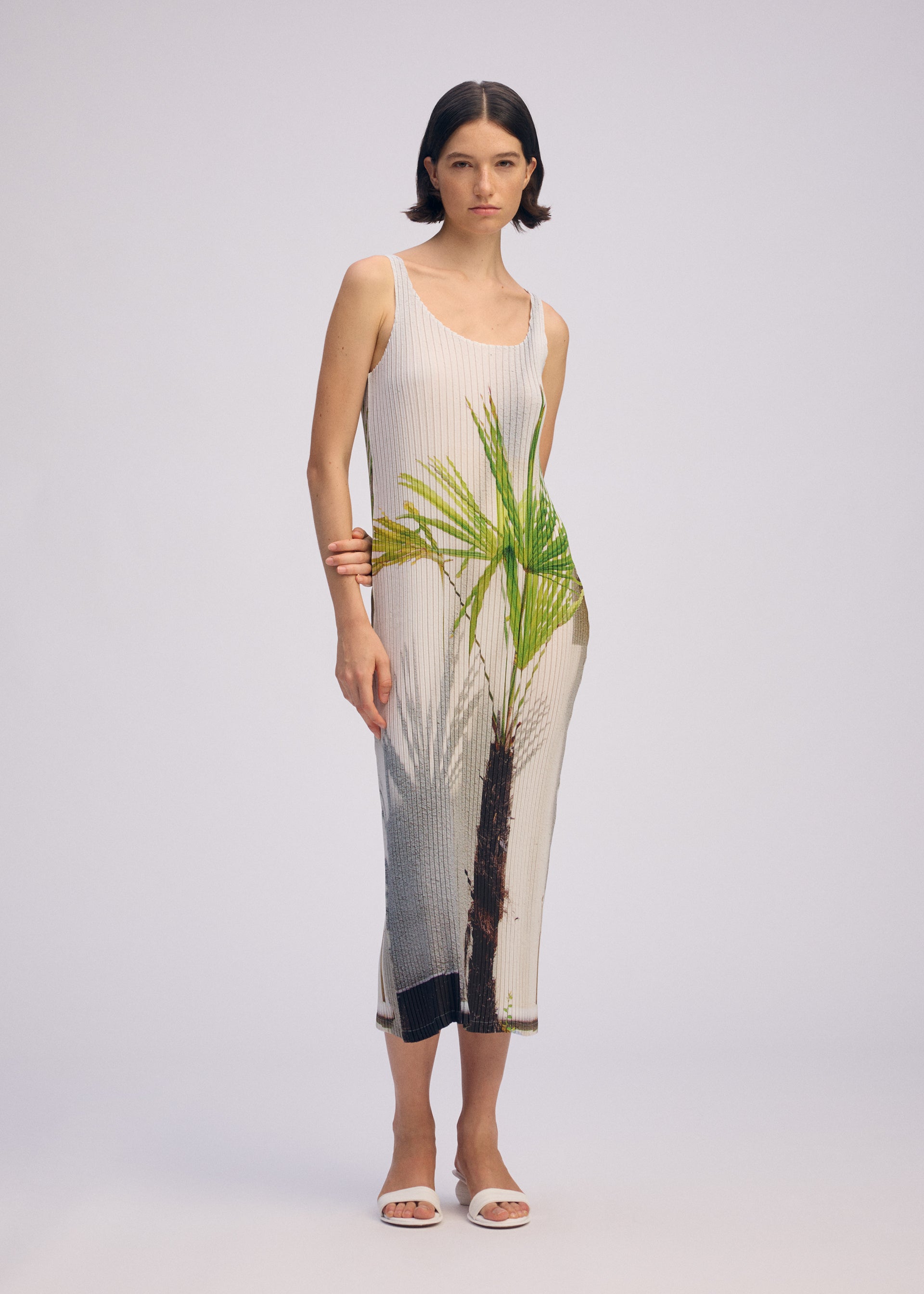 Zoomed view A woman models the ISSEY MIYAKE URBAN JUNGLE PLEATS, a sleeveless, ankle-length dress with bold green palm prints. She pairs it with white sandals, orange bangles, and minimal makeup against a plain backdrop.