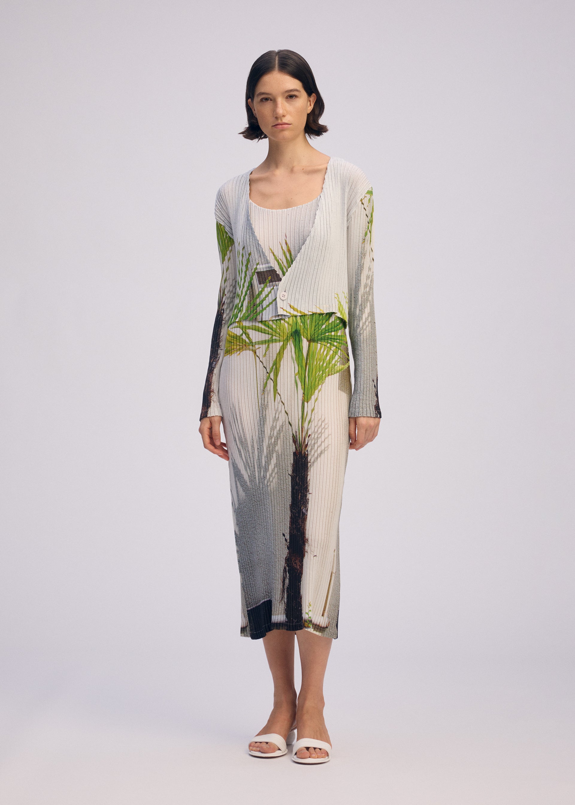Zoomed view A woman with short dark hair wears the ISSEY MIYAKE URBAN JUNGLE PLEATS sleeveless, light-toned pleated dress with a bold jungle print, paired with white open-toe sandals against a plain light background.