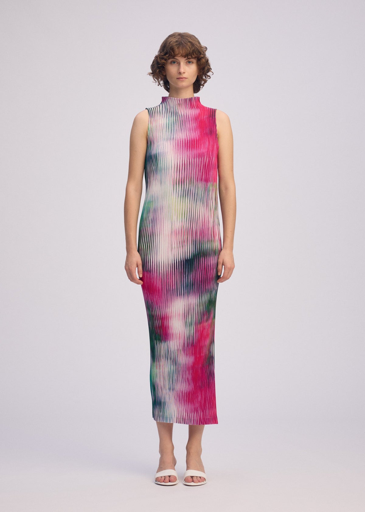A woman faces forward against a plain light background, wearing ISSEY MIYAKE's OBSCURE PLEATS: a high-neck, sleeveless dress with a colorful pink, green, and white abstract pattern, paired with white open-toe heels. view 5 of 7