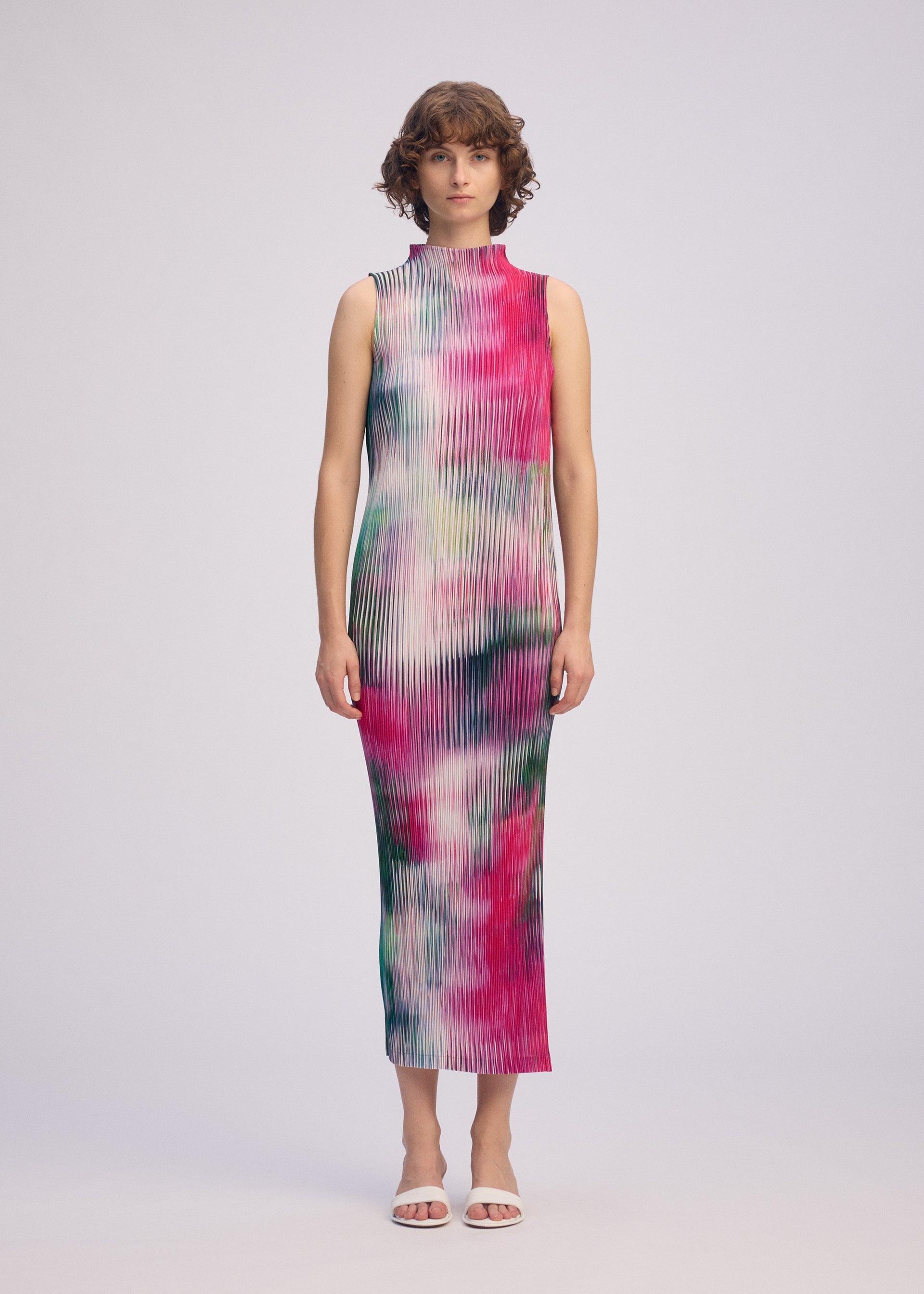 Zoomed view A woman faces forward against a plain light background, wearing ISSEY MIYAKE's OBSCURE PLEATS: a high-neck, sleeveless dress with a colorful pink, green, and white abstract pattern, paired with white open-toe heels.