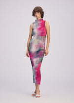 A person wearing the OBSCURE PLEATS high-neck sleeveless pleated polyester dress by ISSEY MIYAKE in vibrant pink, green, and white tie-dye with white sandals stands against a plain light background.