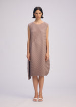 A woman stands facing forward wearing the ISSEY MIYAKE CIRCLE PLEATS PB dress made from plant-derived polyester, with handmade pleats and an asymmetrical hem. She has short dark wavy hair, white sandals, and a plain light background.