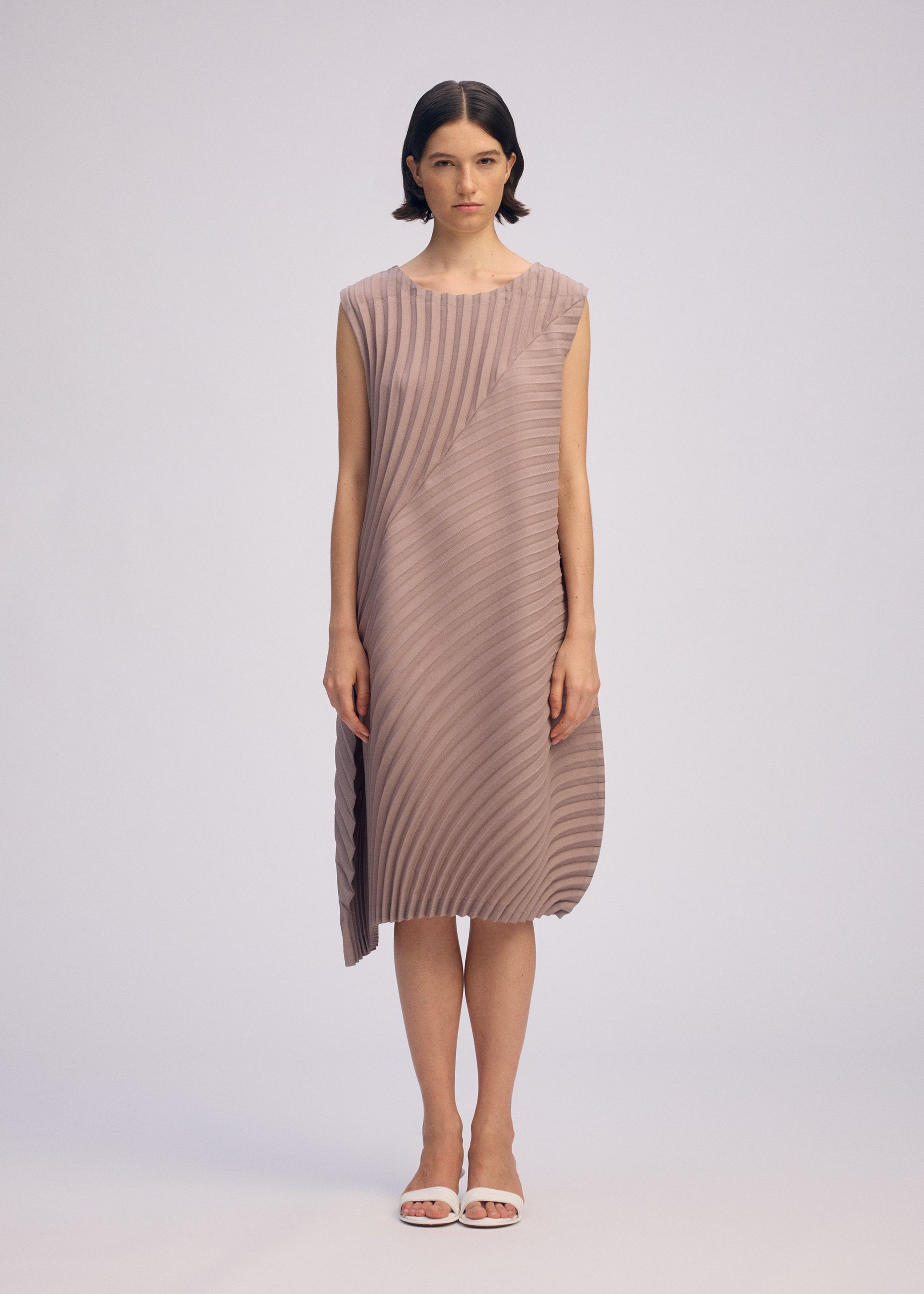 Zoomed view A woman stands facing forward wearing the ISSEY MIYAKE CIRCLE PLEATS PB dress made from plant-derived polyester, with handmade pleats and an asymmetrical hem. She has short dark wavy hair, white sandals, and a plain light background.