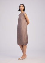 A woman with short dark hair wears the ISSEY MIYAKE CIRCLE PLEATS PB sleeveless taupe knee-length dress with handmade pleats and white open-toe heeled sandals, standing against a plain light background.