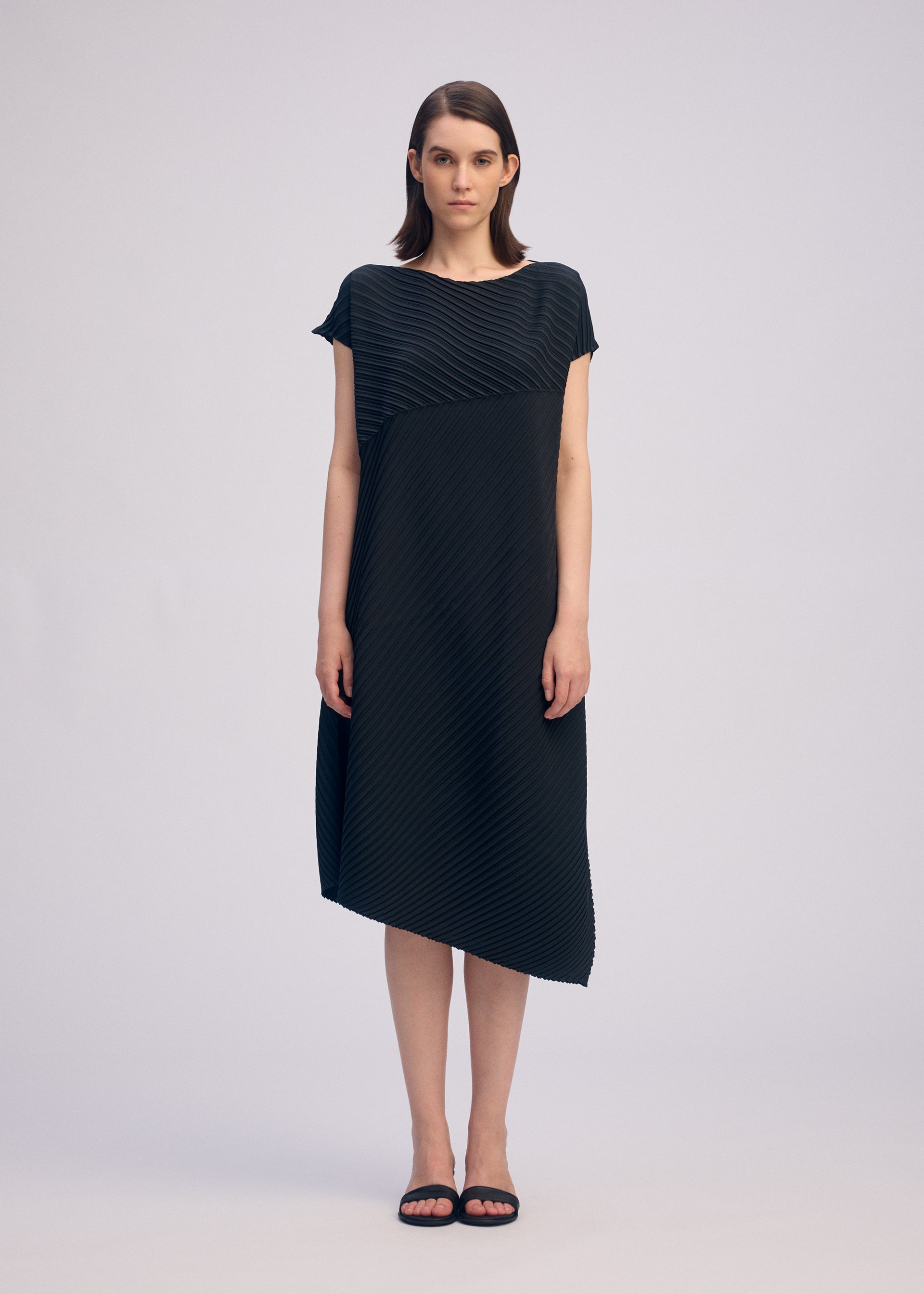 Zoomed view A woman faces forward in the ISSEY MIYAKE PLASTER PLEATS SOLID dress, a loose-fitting black crêpe de Chine style with short sleeves and diagonal pleats. She has straight, shoulder-length hair and wears black sandals against a plain background.