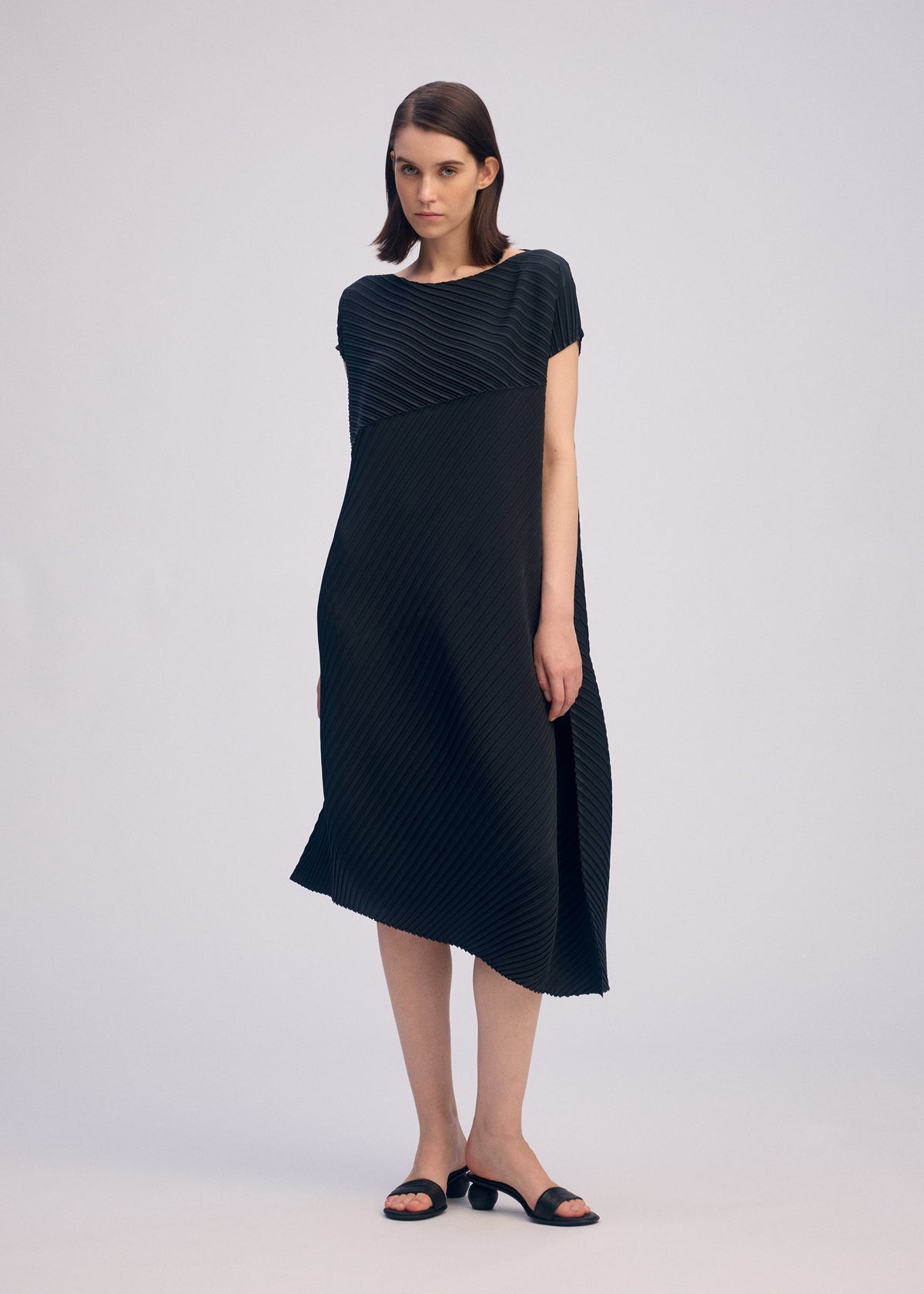 A woman wears the ISSEY MIYAKE PLASTER PLEATS SOLID, a loose-fitting, knee-length black dress with diagonal textured lines. She stands against a light gray background, has straight brown hair, and pairs the look with black sandals. view 6 of 6