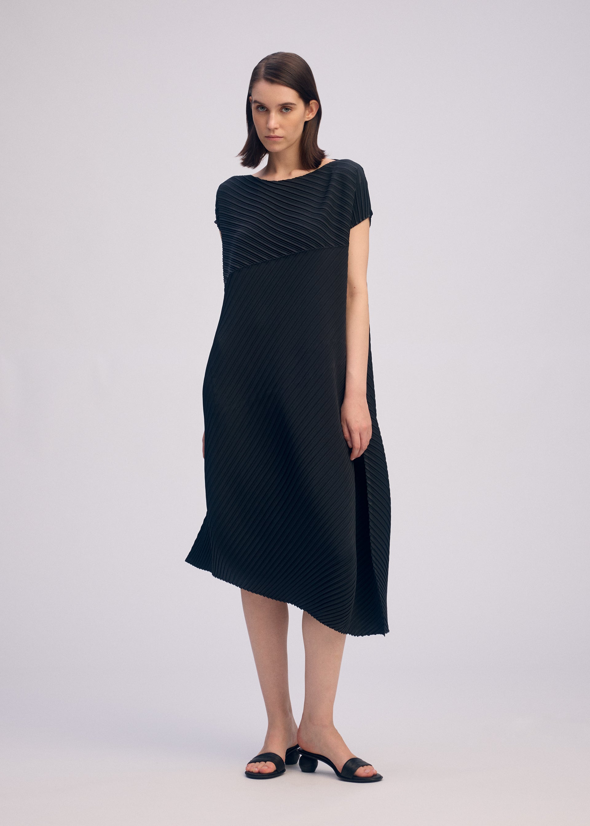 Zoomed view A woman wears the ISSEY MIYAKE PLASTER PLEATS SOLID, a loose-fitting, knee-length black dress with diagonal textured lines. She stands against a light gray background, has straight brown hair, and pairs the look with black sandals.