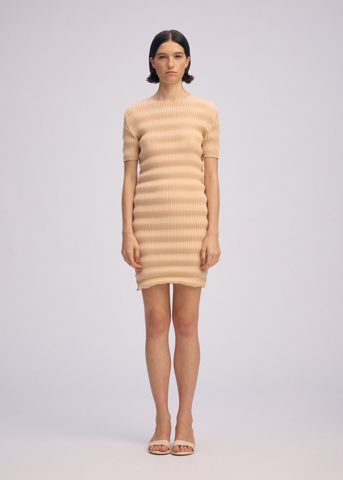 A woman wearing the ISSEY MIYAKE SPRING PLEATS beige ribbed mini dress with handmade pleats and matching open-toe sandals stands against a plain light background, looking straight ahead with a neutral expression. view 4 of 5