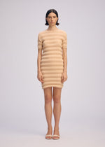 A woman wearing the ISSEY MIYAKE SPRING PLEATS beige ribbed mini dress with handmade pleats and matching open-toe sandals stands against a plain light background, looking straight ahead with a neutral expression.