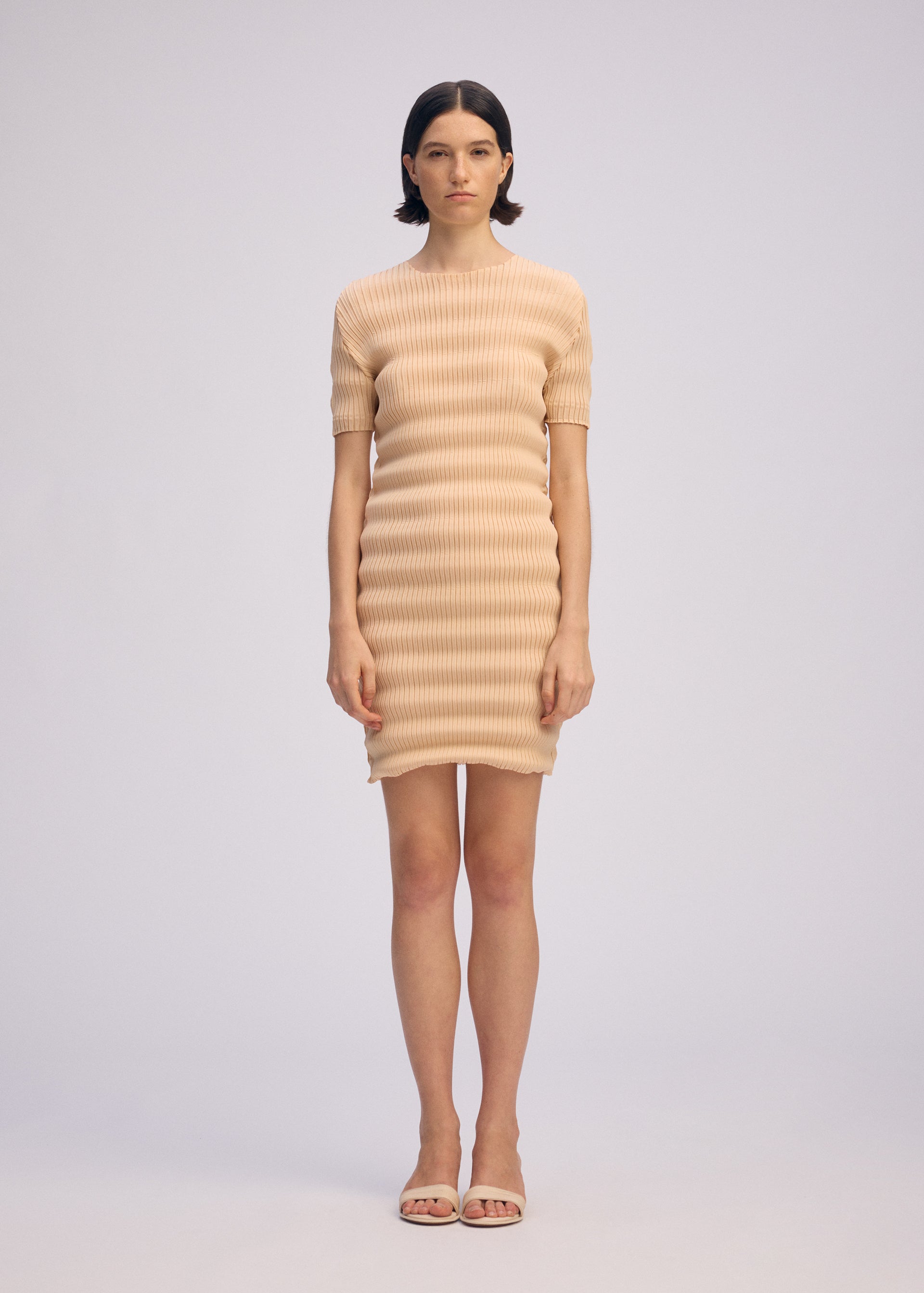 Zoomed view A woman wearing the ISSEY MIYAKE SPRING PLEATS beige ribbed mini dress with handmade pleats and matching open-toe sandals stands against a plain light background, looking straight ahead with a neutral expression.