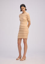 A woman stands against a plain background in the ISSEY MIYAKE SPRING PLEATS beige ribbed mini dress, paired with matching open-toe sandals. Her arms are behind her back as she looks down thoughtfully.