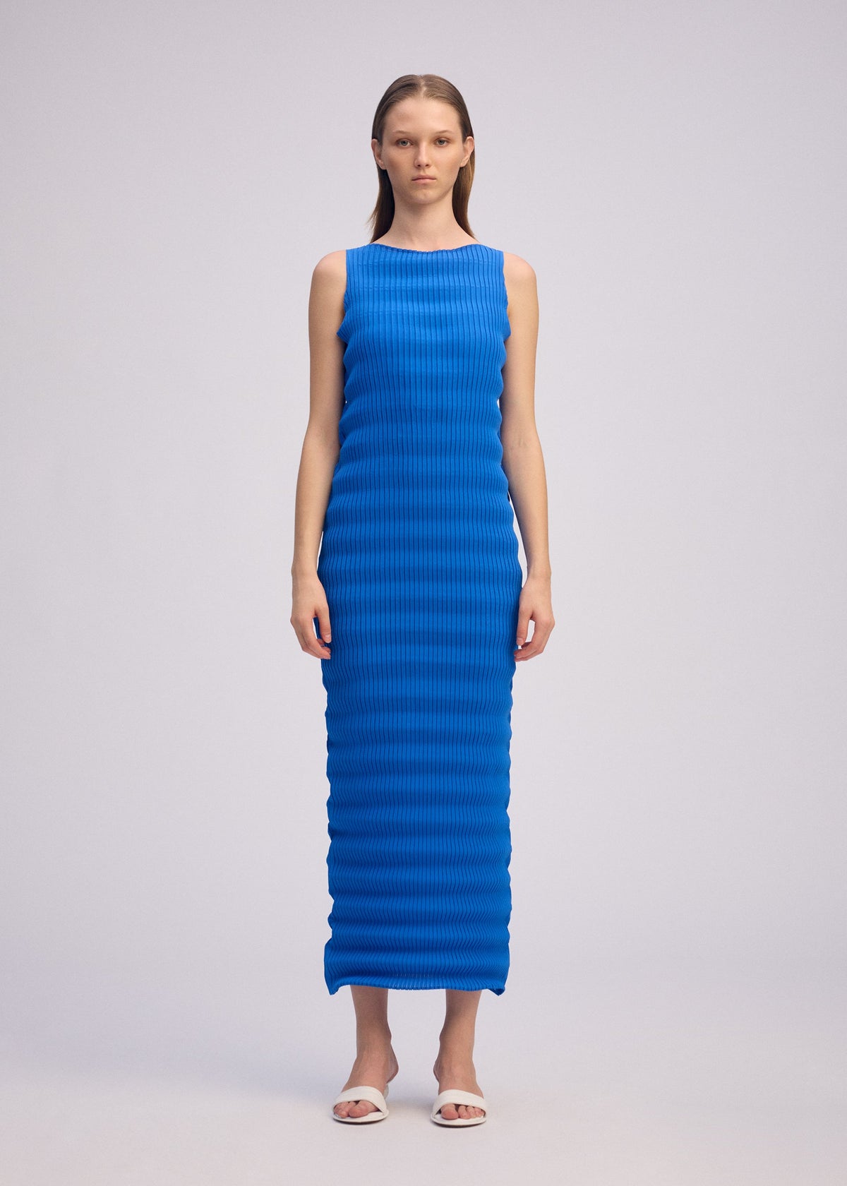 A woman faces forward in the ISSEY MIYAKE SPRING PLEATS sleeveless, bright blue, ankle-length dress with a ribbed texture, paired with white sandals against a plain light gray background. view 4 of 5